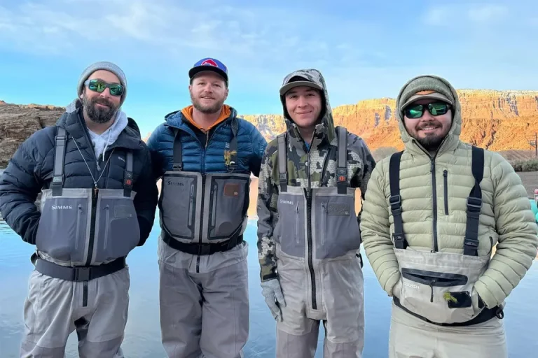 Four About Trout fly fishing guides in waders standing in shallow river water at the base of a red sandstone canyon at sunrise