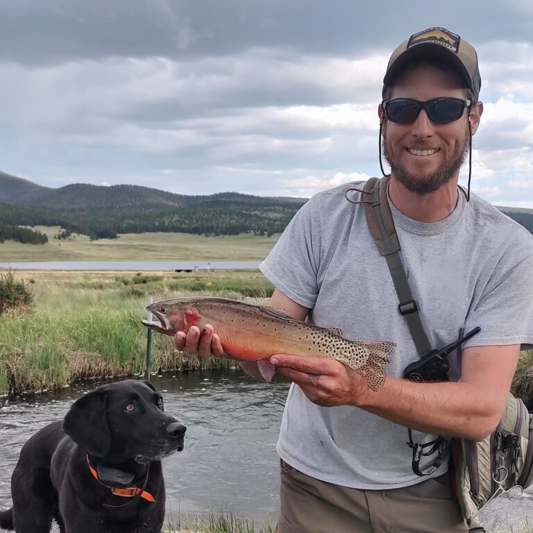 Bryan Ferguson holding a Rio Grande cutthroat trout streamside, his black Lab beside him and a reservoir in the background.