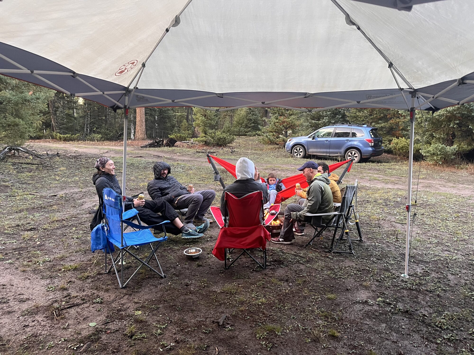 Volunteers sitting in camp chairs under a canopy at a forest campsite.